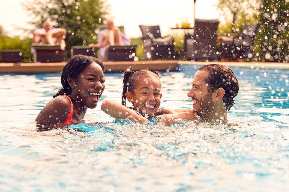 Smiling Family On Summer Holiday Having Fun Splashing In Outdoor Swimming Pool